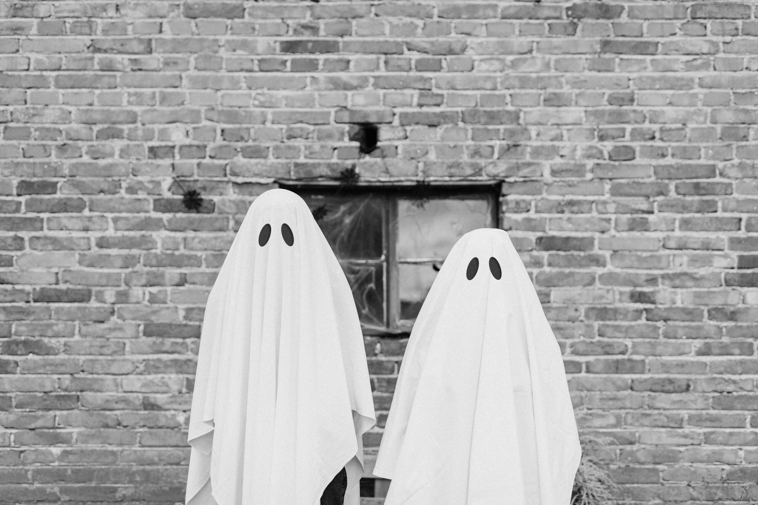Two figures covered in bedsheets to look like ghosts stending in front of a brick wall